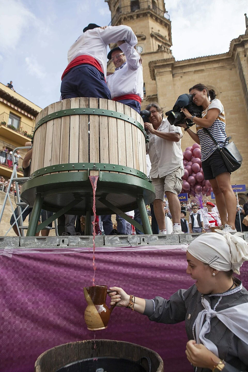 Two men in a barrel crushing grapes with their feet and a woman pouring this from the barrel into a jug